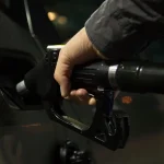 Driver checking the owner’s manual and fuel door label beside a sedan at a gas station